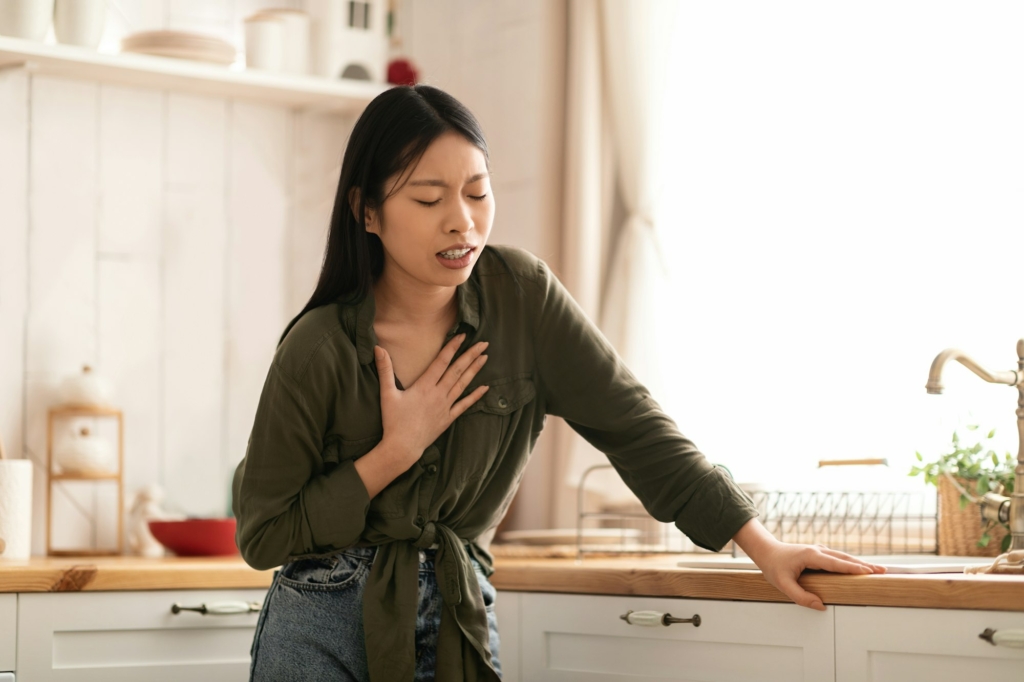 Sick asian woman suffering from heartburn in kitchen, touching chest, caused by hiatal hernia
